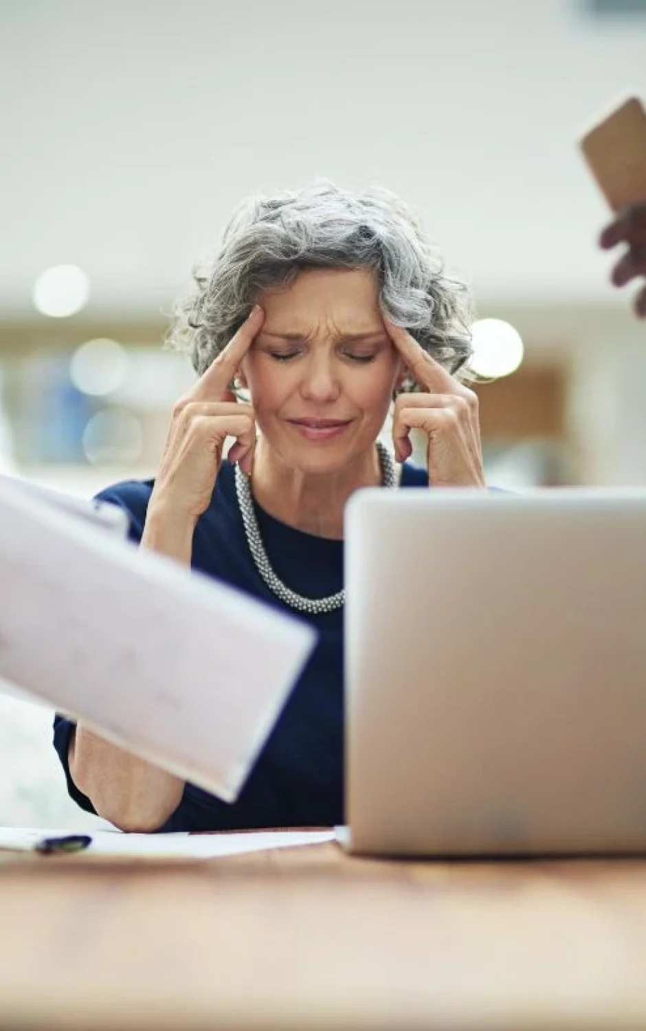 An older woman with gray hair, seated at a desk with a laptop, looks stressed and rubs her temples. She is surrounded by two people holding documents and a smartphone, all vying for her attention. The background is blurred, indicating a busy office environment.