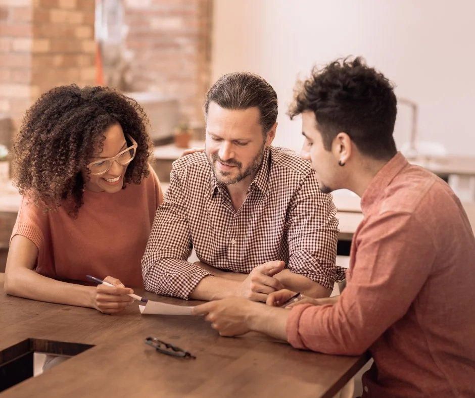 Three people sit at a table in a well-lit room, collaborating on a document. The woman on the left is smiling and holding a pen, the man in the middle is attentively looking at the paper, and the man on the right is explaining something. They all look focused and engaged.