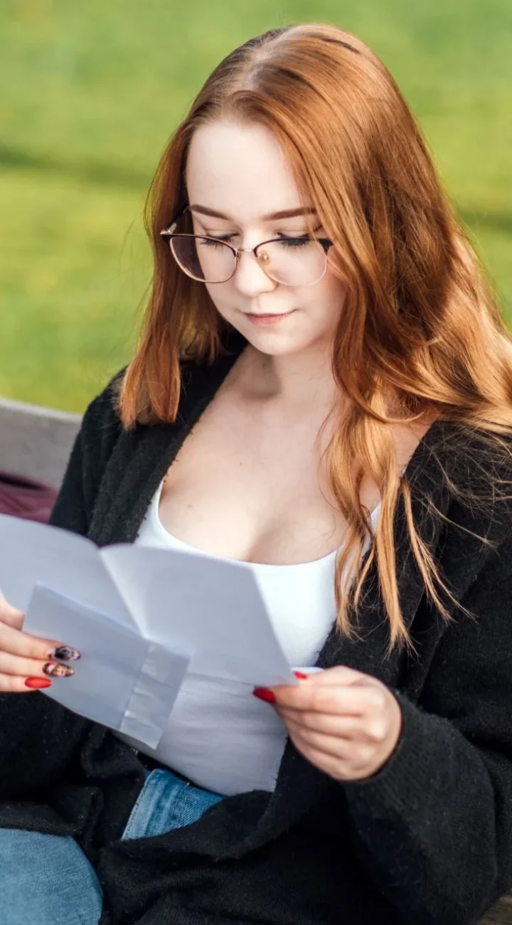 A woman with long, reddish-brown hair and glasses is sitting outdoors, reading a piece of paper. She is wearing a white top and a black cardigan. The background is green, suggesting she is in a park or garden.