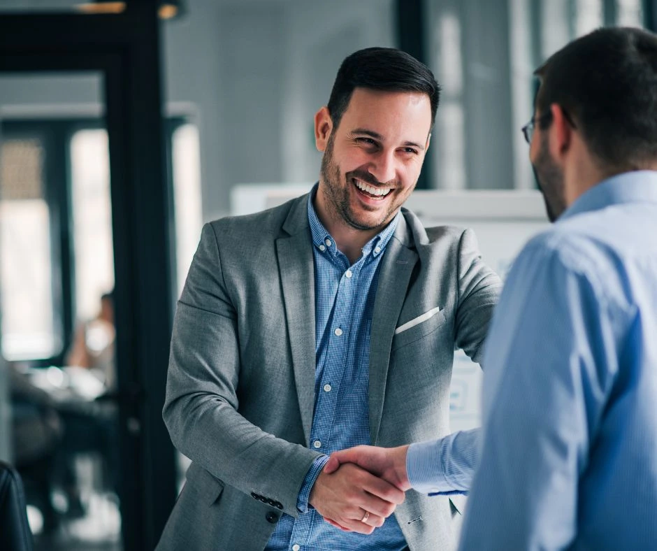 Two men are shaking hands and smiling in an office setting. The man on the left is wearing a grey blazer over a blue shirt, while the man on the right is in a light blue dress shirt. They appear to be engaged in a positive conversation.