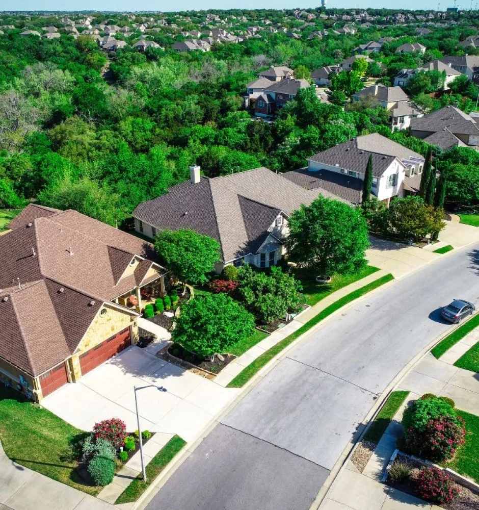 Aerial view of a suburban neighborhood with large houses surrounded by greenery and trees. The homes have well-maintained lawns and are situated along a curving road. The neighborhood is bordered by a densely wooded area in the background.