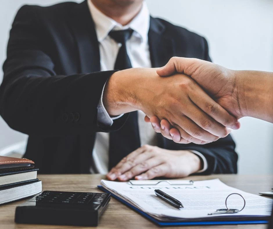 Two people are shaking hands over a desk, with documents, a calculator, and a pen organized neatly on top. One person is wearing a suit and tie, while the other person's hand is visible. The scene appears professional, suggesting a business agreement.