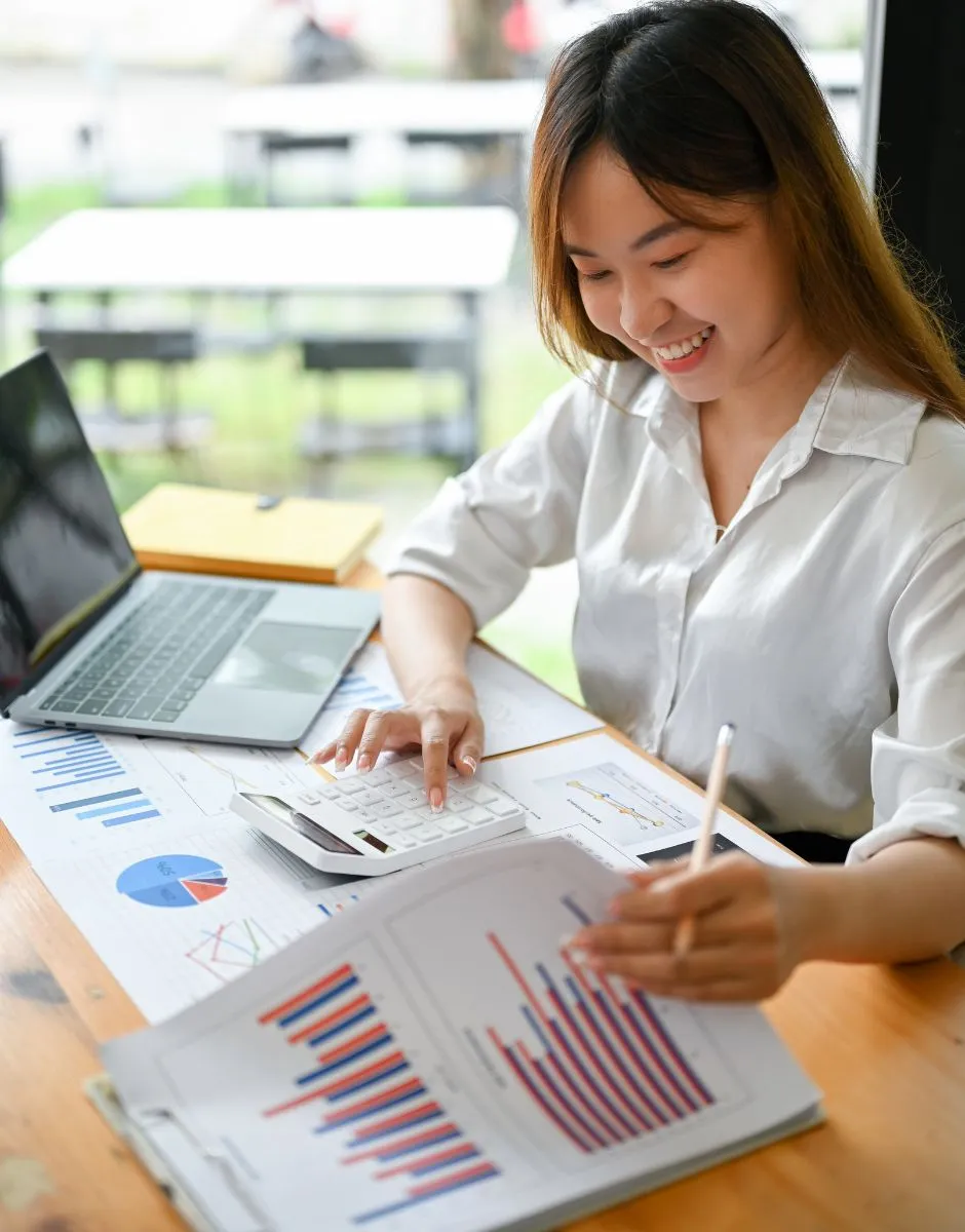 A woman in a white shirt is sitting at a desk and smiling as she works with charts and graphs. She is holding a paper with a bar chart and using a calculator. A laptop, notebooks, and more papers with graphs are spread out on the desk in front of her.